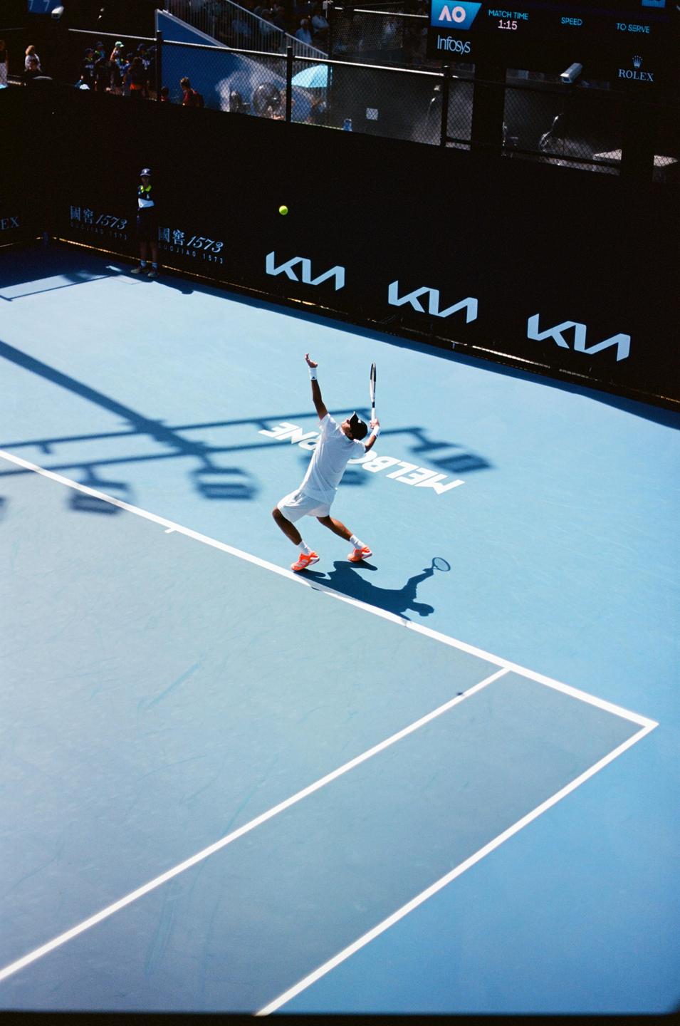 A tennis player in white serves a tennis ball on a blue court.