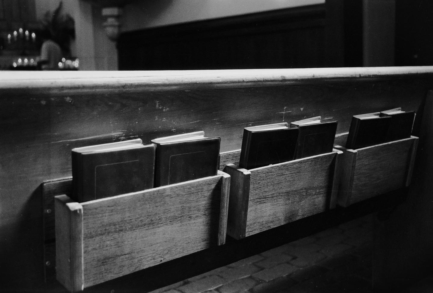 Several books are stored in wooden holders on the back of a church pew.
