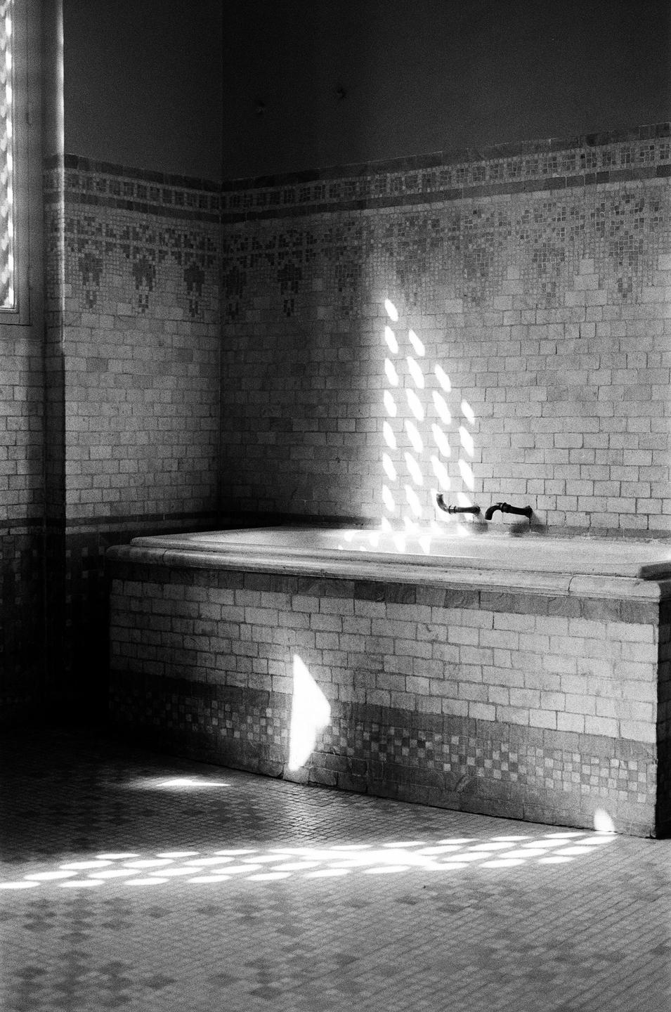 Sunlight streams through a window, casting patterns on the tiled walls and floor of a bathroom with a large bathtub.
