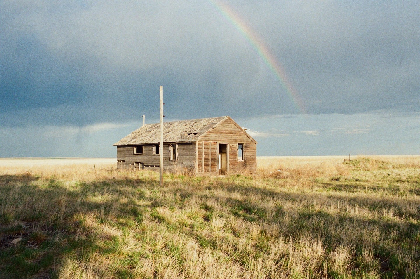 An abandoned wooden house stands in a field under a cloudy sky with a rainbow.