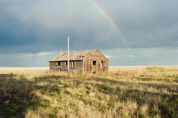 An abandoned wooden house stands in a field under a cloudy sky with a rainbow.