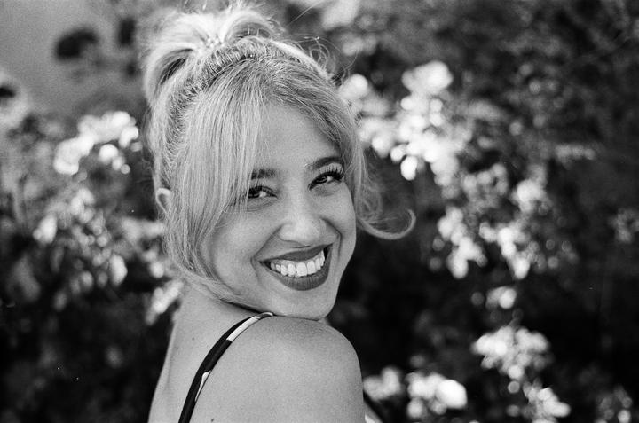 A woman with her hair in a bun and bangs smiles at the camera with foliage in the background.