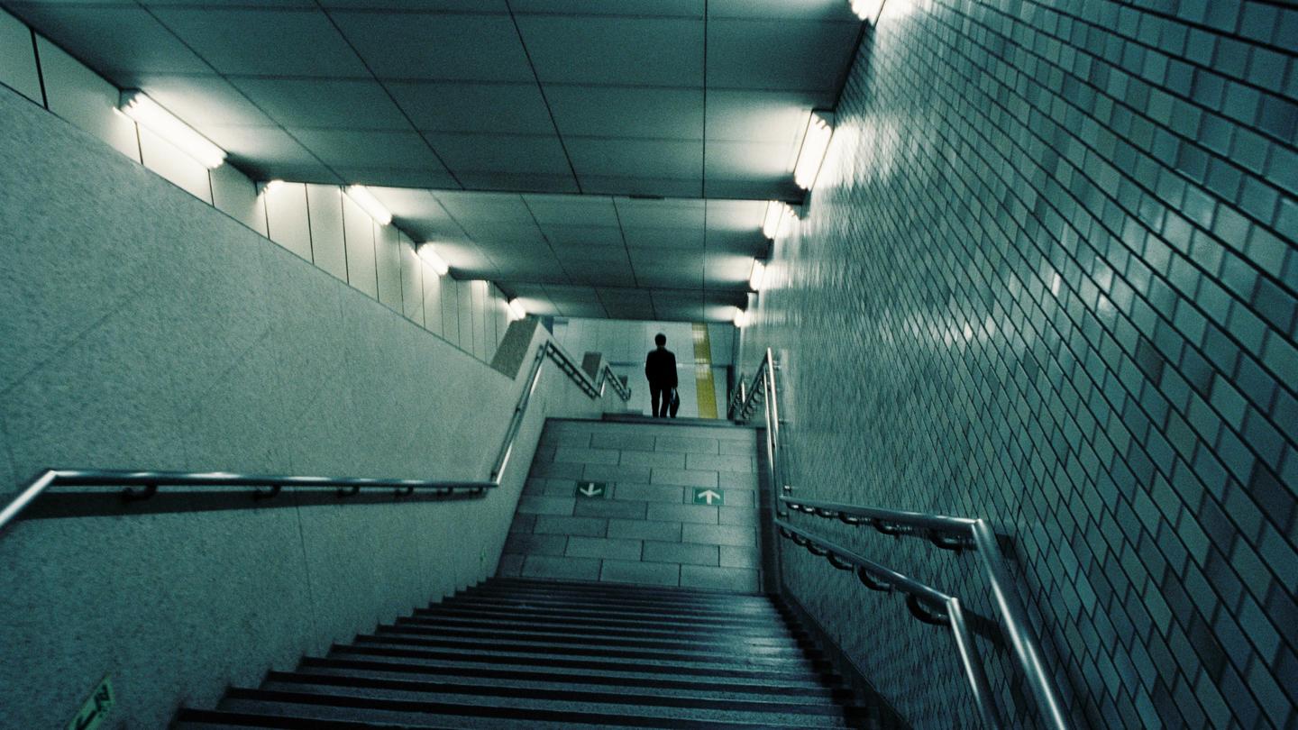 A man stands at the bottom of a subway staircase with fluorescent lights illuminating the walls.