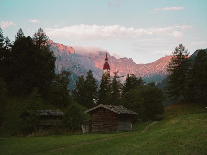 A small wooden cabin sits in a grassy field with a church steeple and mountains in the background.