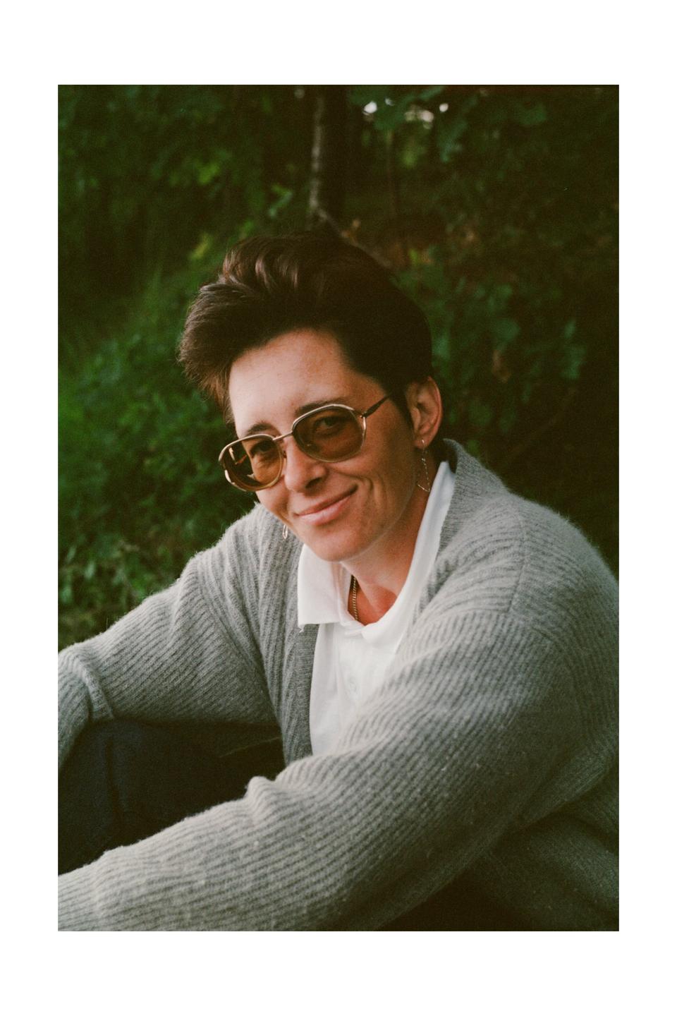 A woman wearing sunglasses and a cardigan smiles at the viewer while sitting outdoors.