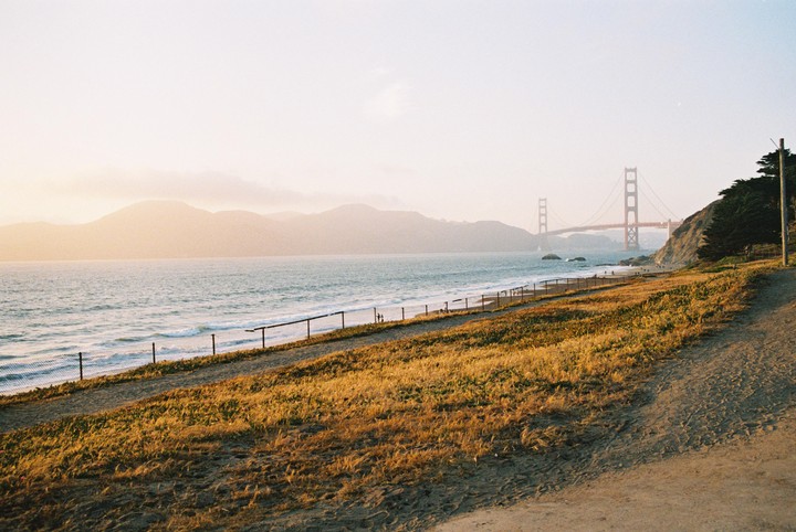 The Golden Gate Bridge is visible in the distance across a body of water, with a grassy and sandy area in the foreground.