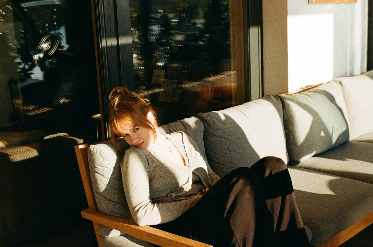 A woman with red hair sits on a sofa bathed in sunlight.