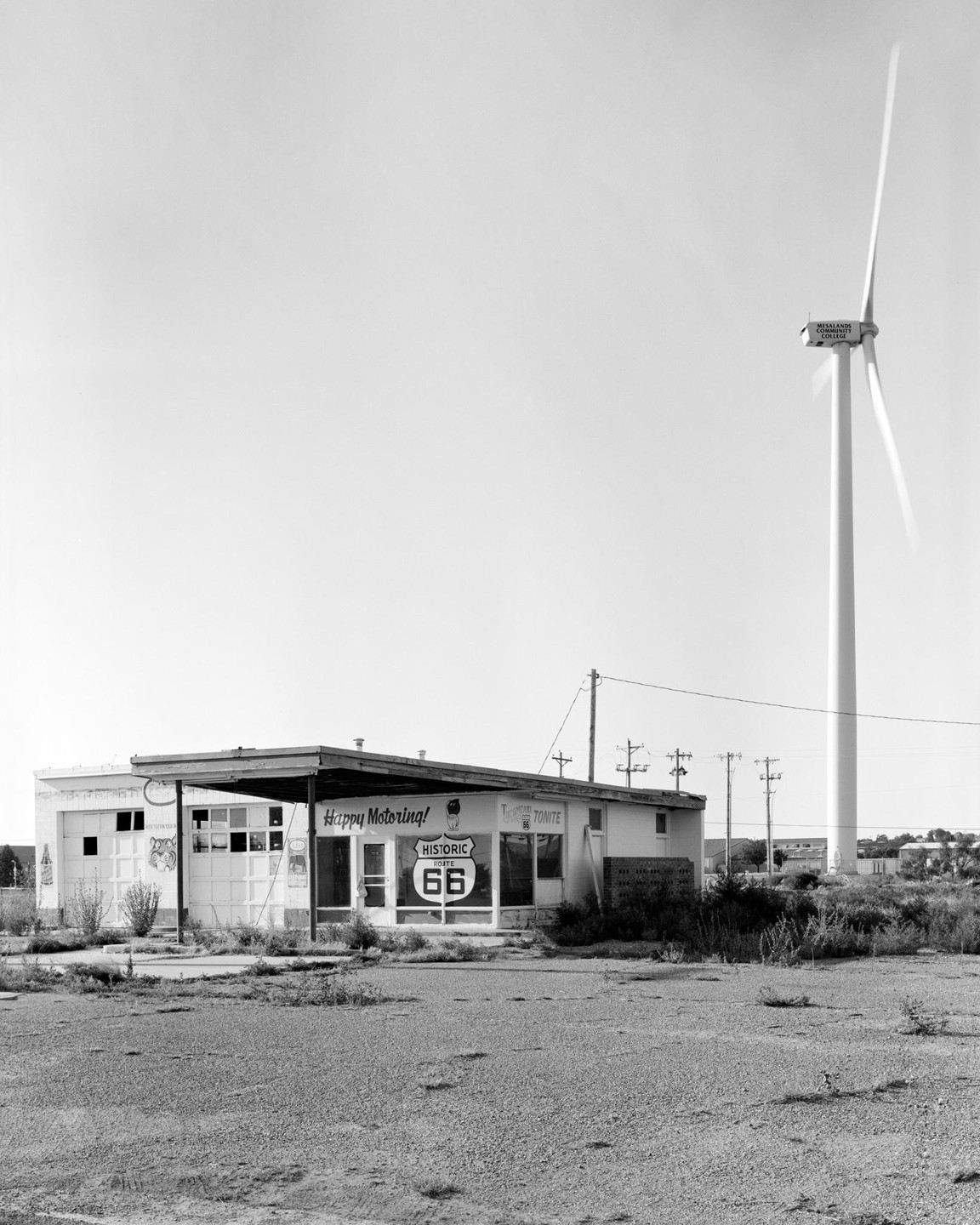 An abandoned gas station with a "Historic Route 66" sign on its window stands next to a large wind turbine under a clear sky.
