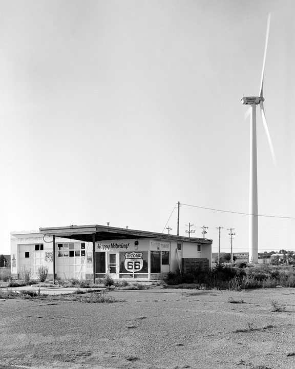 An abandoned gas station with a "Historic Route 66" sign on its window stands next to a large wind turbine under a clear sky.
