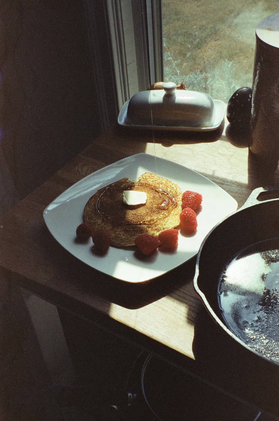 A pancake with butter and raspberries is on a white plate next to a cast iron pan and a butter dish.