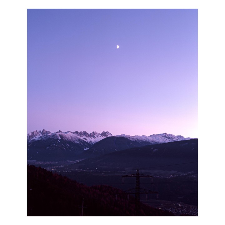 A crescent moon hangs in a purple sky above snow-capped mountains and a valley with a power line.