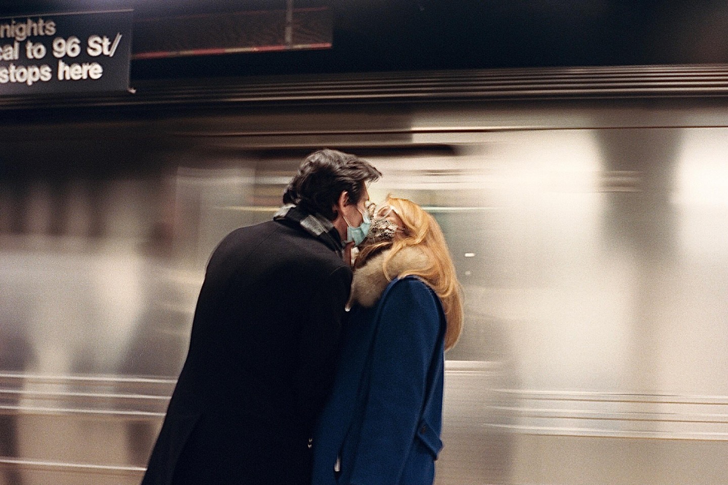 A couple wearing face masks kisses in a subway station as a train passes by.