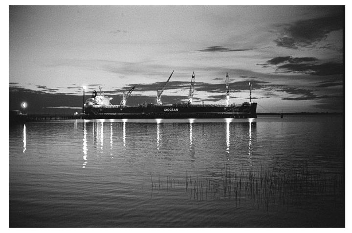 A large cargo ship with cranes is docked at a pier, reflecting its lights on the water under a cloudy sky.