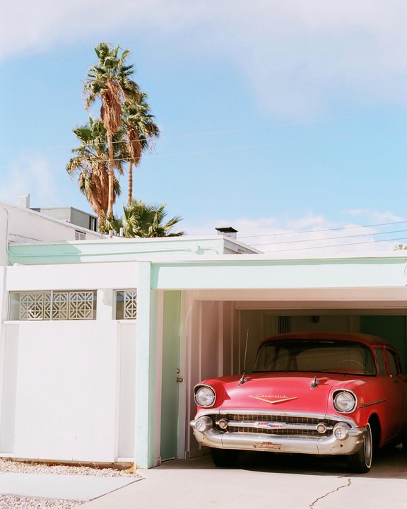 A red vintage car is parked in a garage with palm trees and a blue sky in the background.