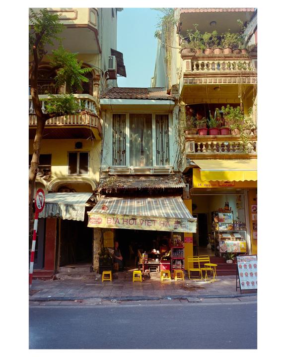 A street in Hanoi is lined with buildings, one of which has a striped awning over a storefront with a person sitting inside.