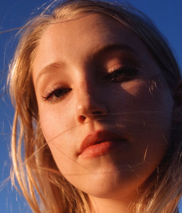 A woman's face is illuminated by golden light against a blue sky, with some hair strands blowing across her face.