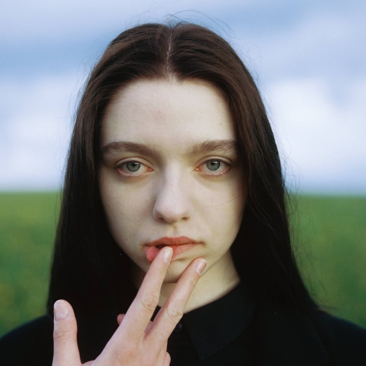 A woman with long dark hair looks directly at the viewer, with her fingers touching her lips.