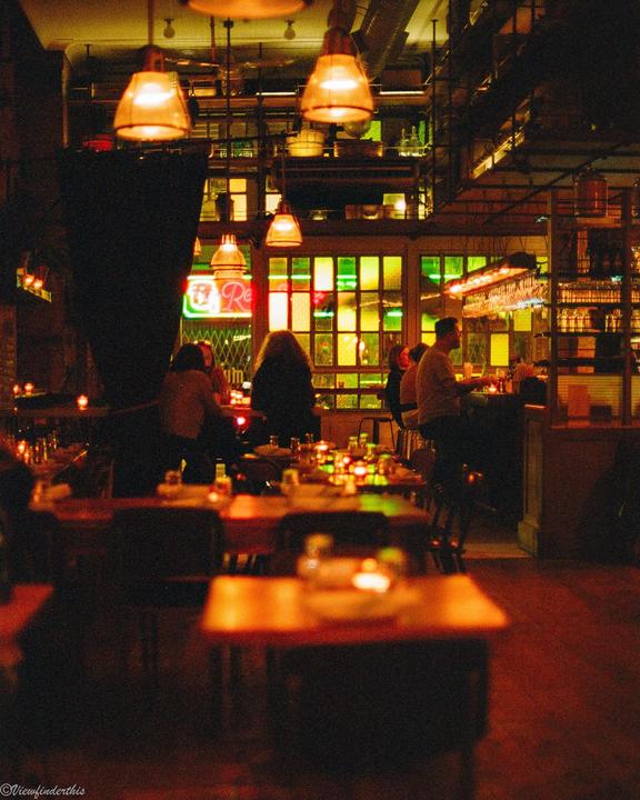 People are gathered in a dimly lit restaurant with warm lighting and a neon sign visible through a window.