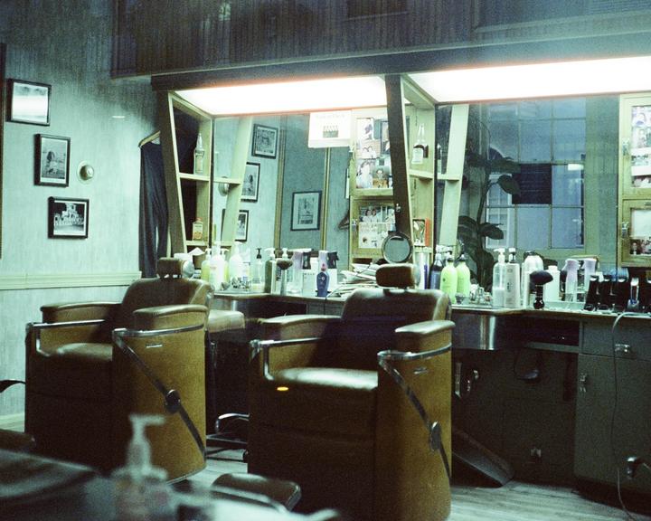 Two empty barber chairs face a mirror and counter filled with hair products in a barbershop at night.