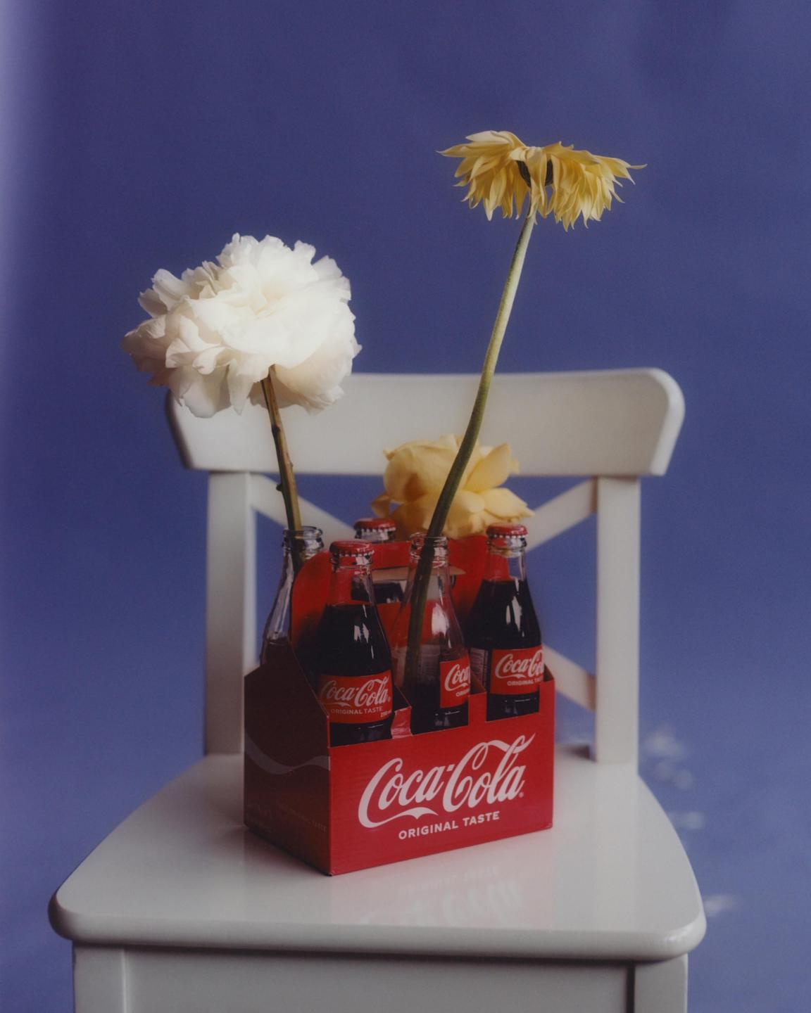 A six-pack of Coca-Cola bottles with flowers in them sits on a white chair against a blue background.