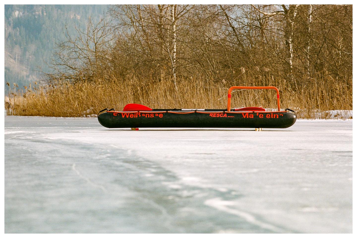 A black rescue sled with orange lettering and a red paddle rests on a frozen lake with reeds and trees in the background.