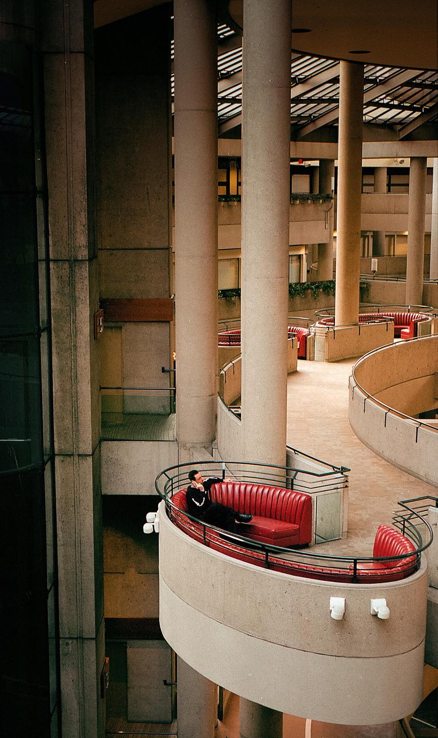A man reclines on a red sofa on a curved balcony in a brutalist building.