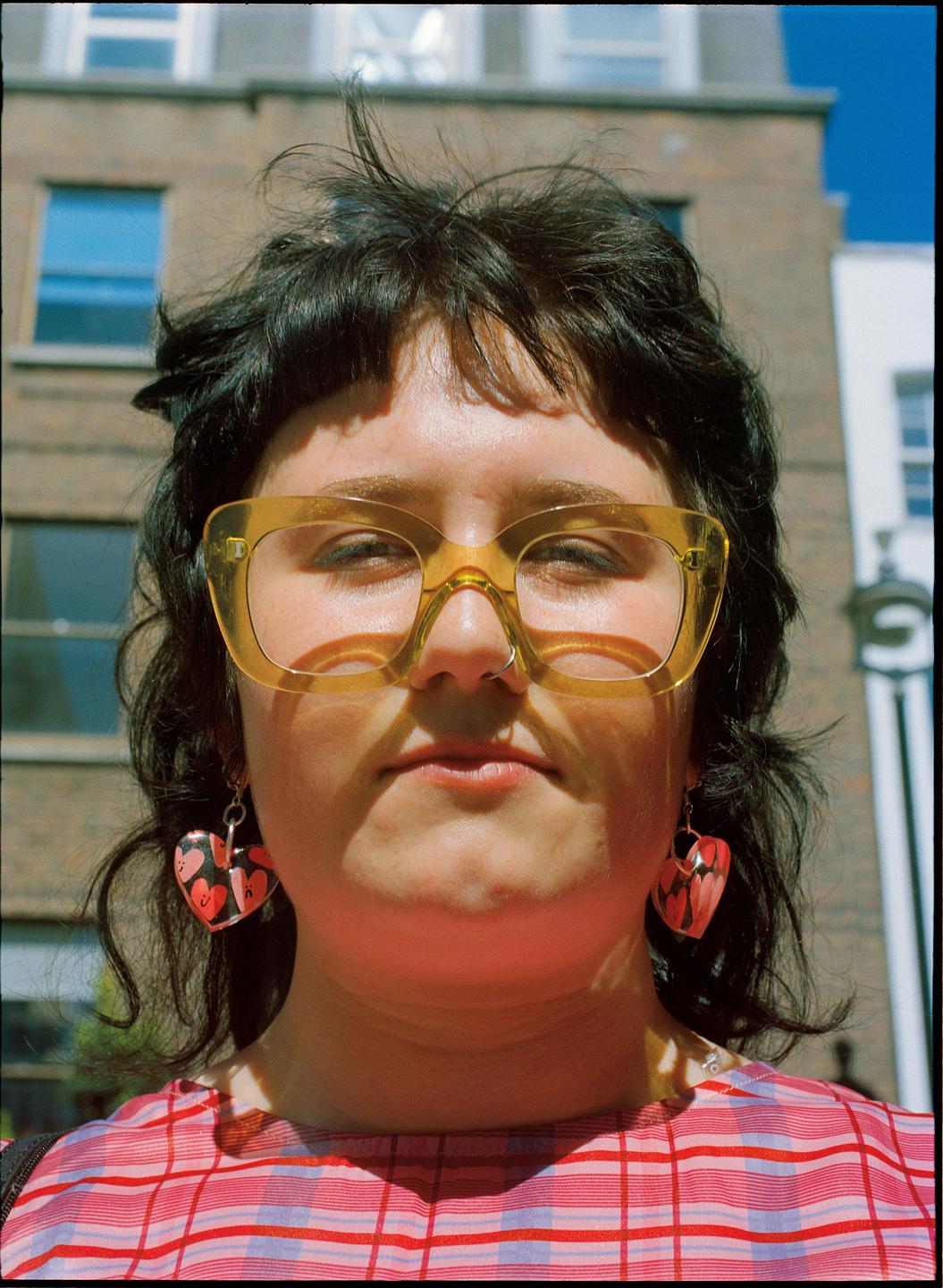 A woman with yellow glasses and heart earrings looks directly at the viewer.