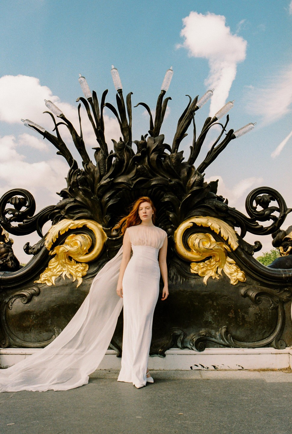 A woman in a white dress with a long train stands in front of an ornate black and gold sculpture under a blue sky with clouds.