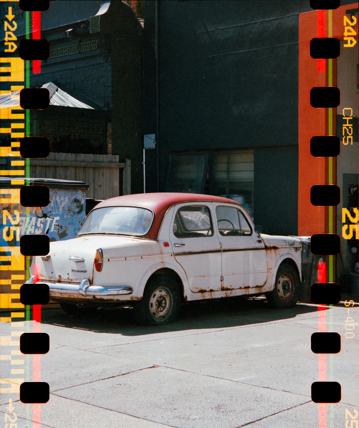 A rusty white car with a red roof is parked on a paved surface next to a dark building and a dumpster.