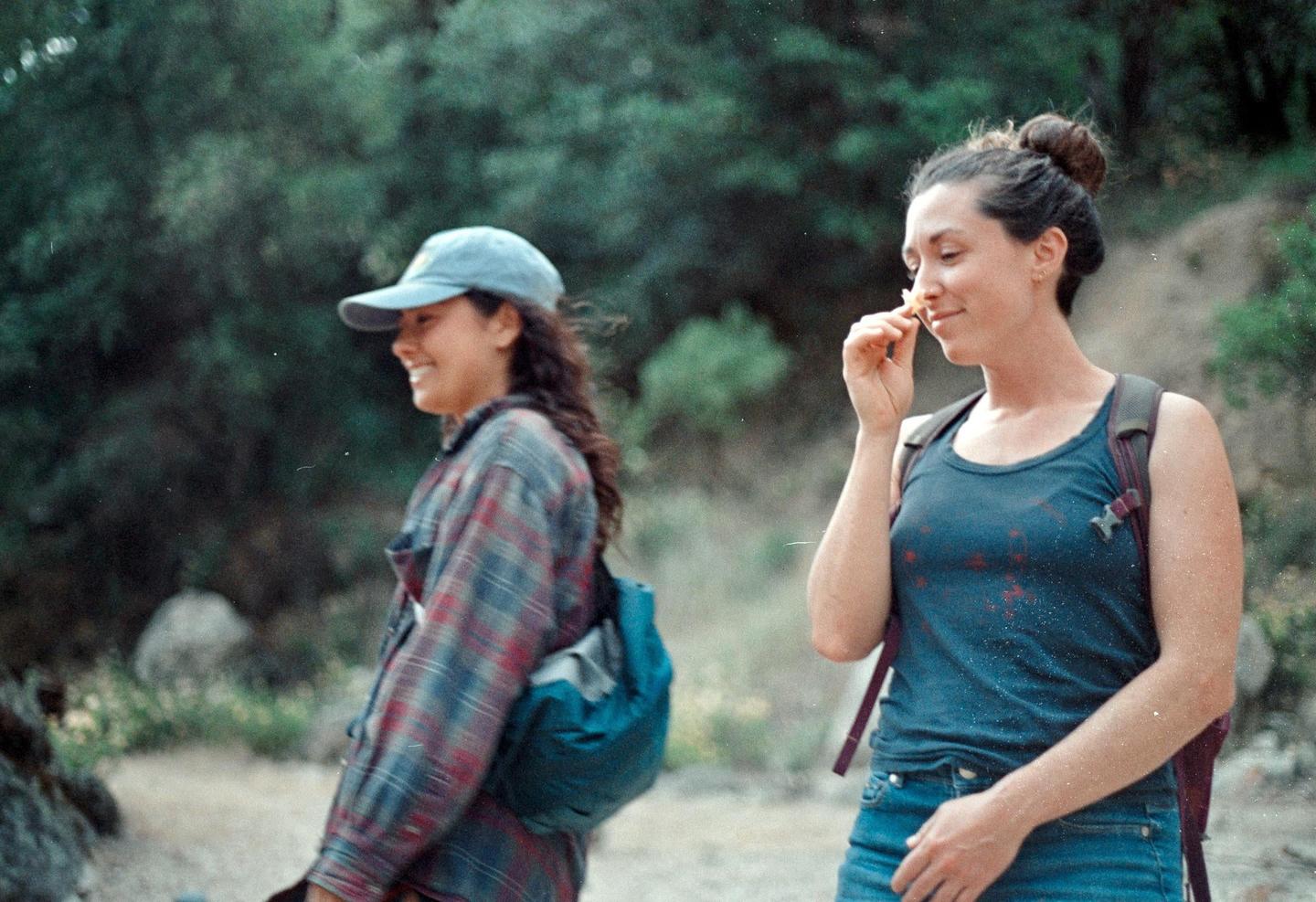 Two women are walking outdoors, one smiling and the other smelling a small flower.