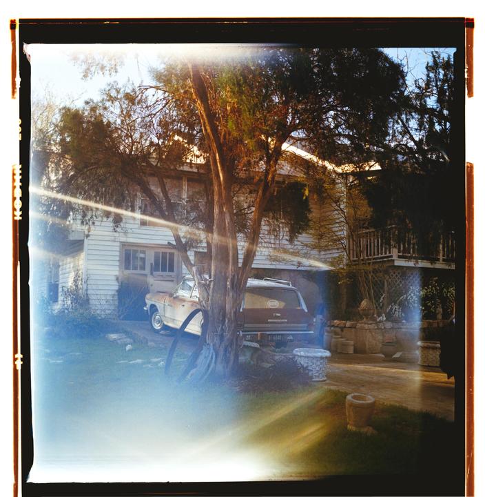 A white house with a porch and a car in the driveway is partially obscured by a tree and lens flare.