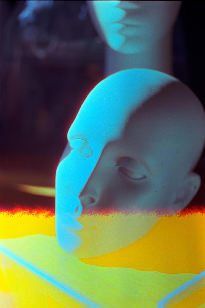 A mannequin head with closed eyes is illuminated by blue light, resting on a yellow surface with a second mannequin head visible in the background.
