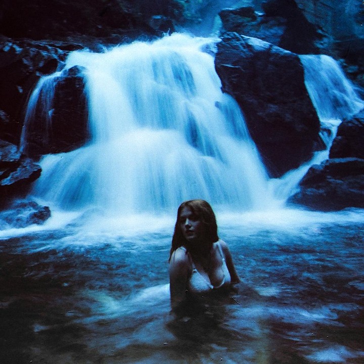 A woman in a white top is in the water in front of a waterfall.
