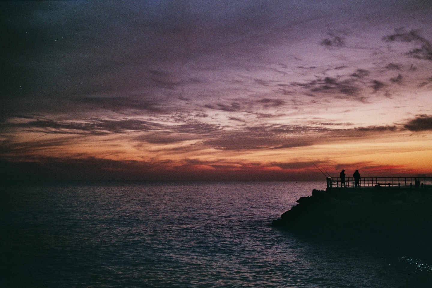 Two men are fishing from a pier at sunset.