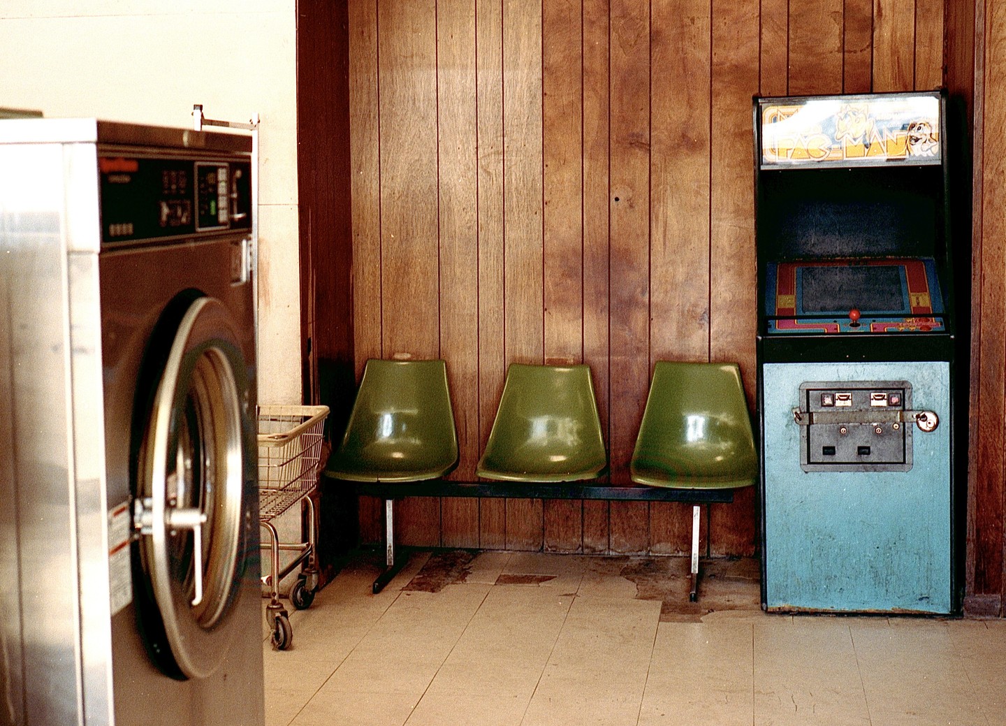 A laundromat interior features a washing machine, a row of three green chairs, and an arcade game against a wood-paneled wall.