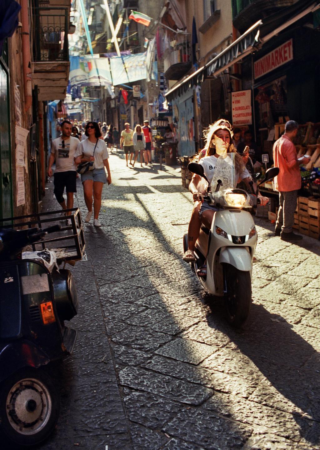 A woman rides a scooter down a narrow, crowded street with buildings and shops on either side, with sunlight and shadow dappling the cobblestone street.