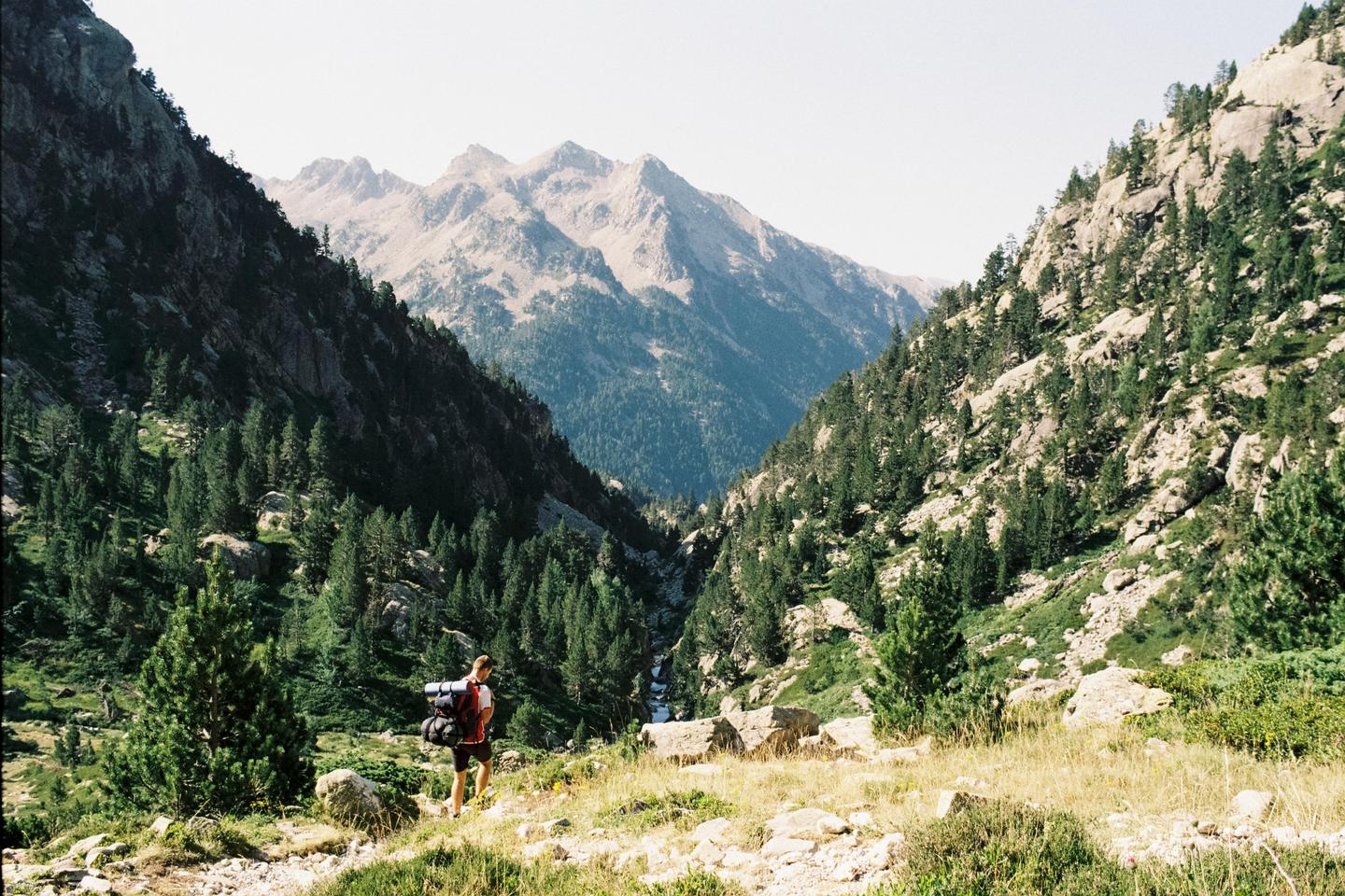 A man with a backpack stands on a grassy hill overlooking a mountain valley with a river.