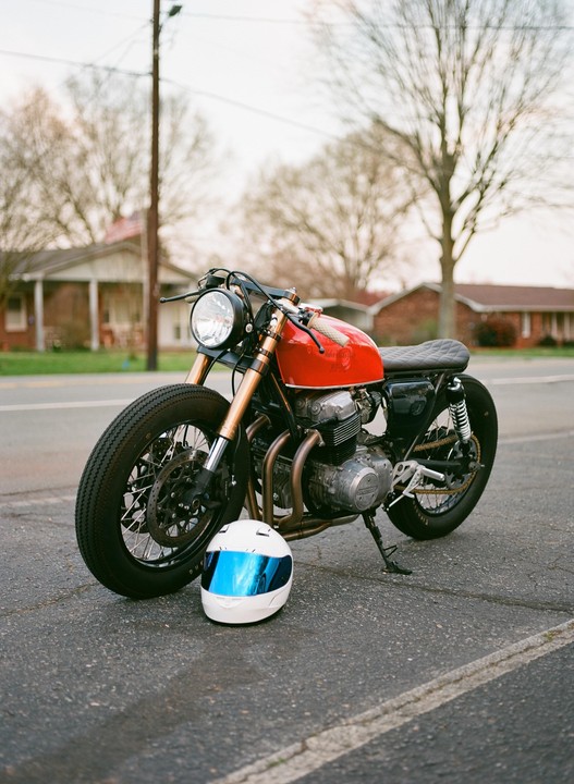 A red motorcycle with a white helmet in front of it is parked on an asphalt road with houses and trees in the background.
