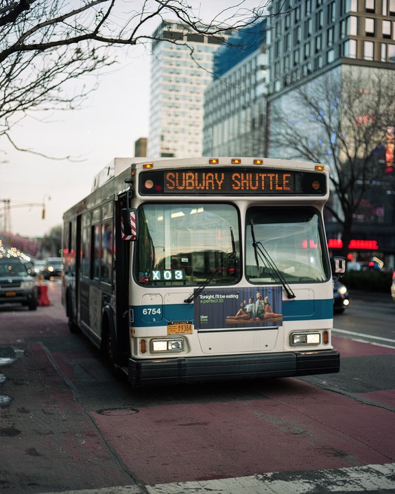 A subway shuttle bus is parked on a city street with tall buildings in the background.