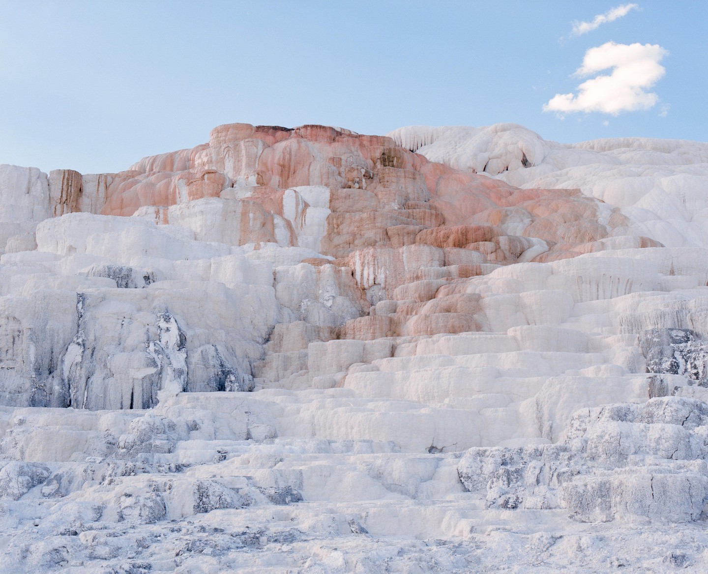 Terraces of white and orange travertine are visible under a blue sky with a single cloud.
