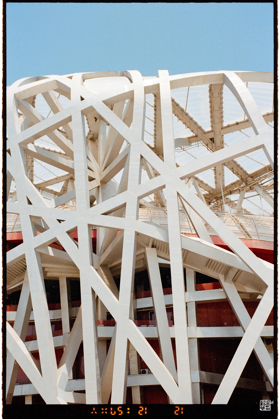 A close-up view of the white, crisscrossing beams of the Beijing National Stadium is shown against a clear blue sky.