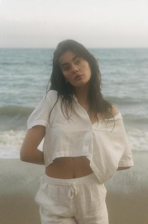 A woman with wet hair and a white shirt stands on a beach with the ocean in the background.