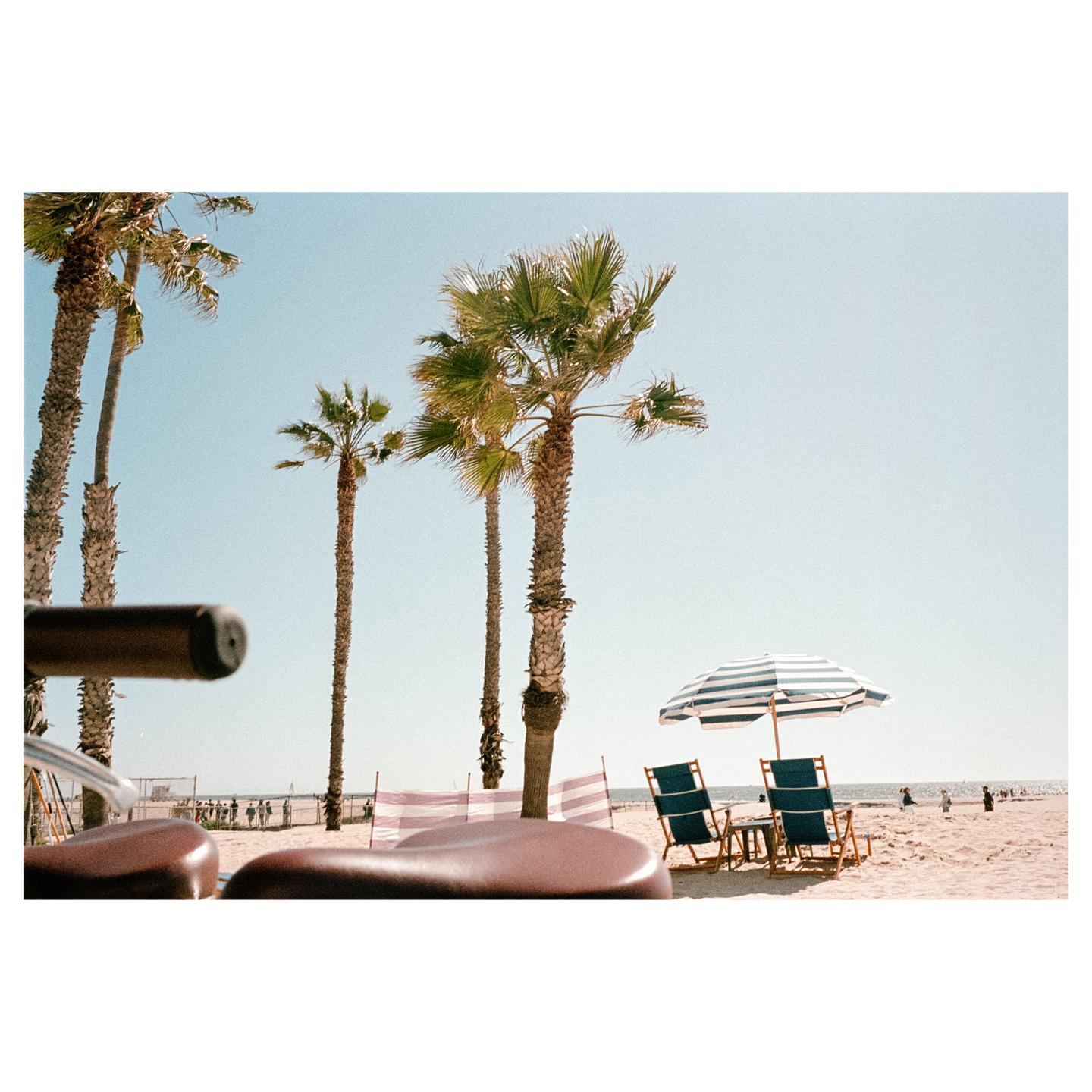 A beach scene features palm trees, a striped umbrella, and two beach chairs on the sand, with a bicycle seat in the foreground.