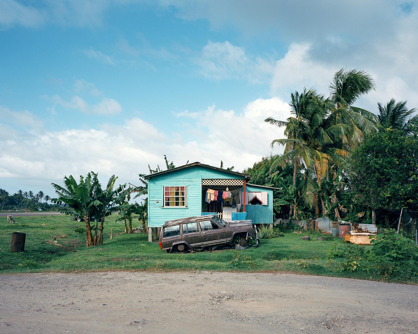 A turquoise house with laundry hanging on the porch stands next to a broken-down car and palm trees under a cloudy sky.