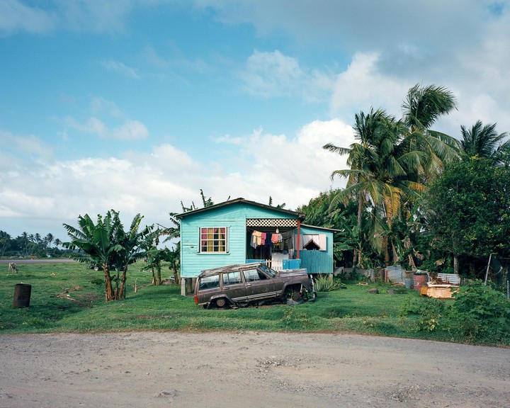 A turquoise house with laundry hanging on the porch stands next to a broken-down car and palm trees under a cloudy sky.