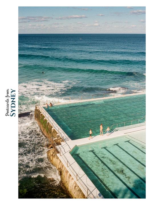 A swimming pool built into a rocky coastline is filled with clear blue water, with the ocean stretching out to the horizon under a partly cloudy sky.