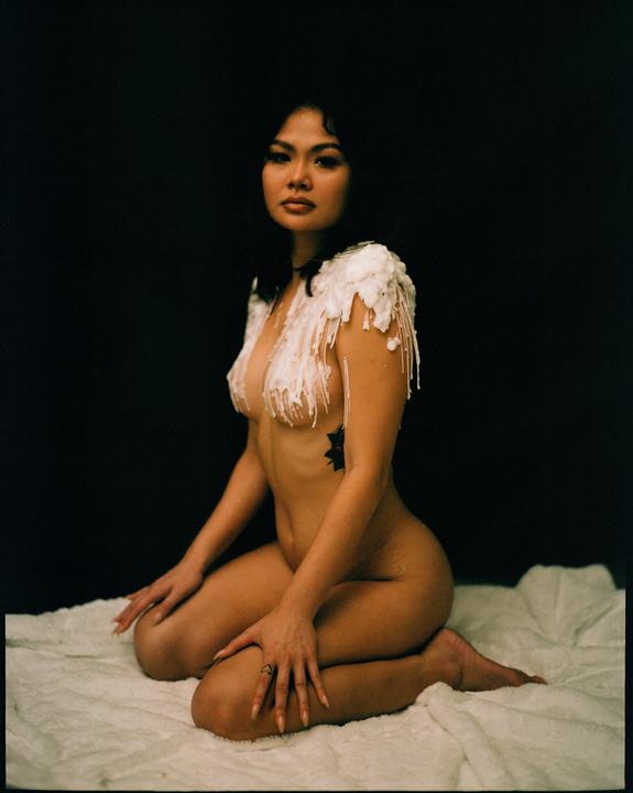 A woman kneels on a white blanket with white wax dripping from her shoulders and chest.