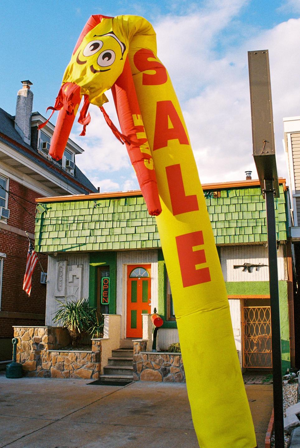 A yellow inflatable tube man with "SALE" written on it stands in front of a building with a green roof and an orange door.