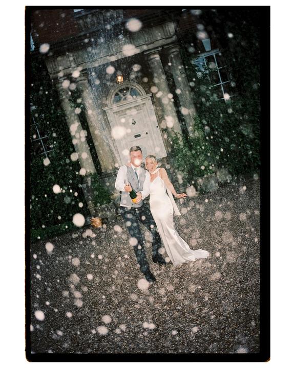 A couple stands in front of a large house with champagne spraying around them.