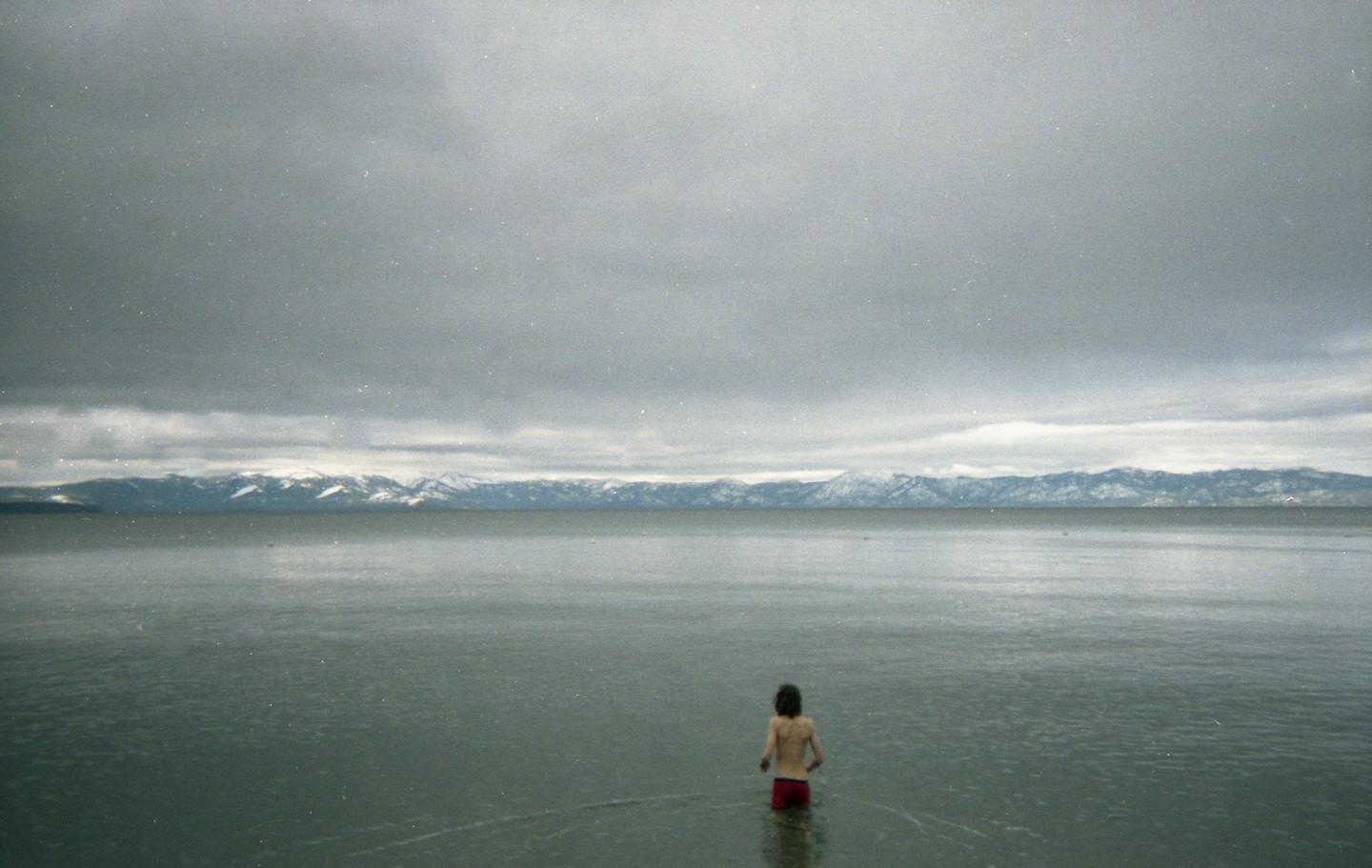 A person stands in a vast lake with snow-capped mountains in the distance under a cloudy sky.